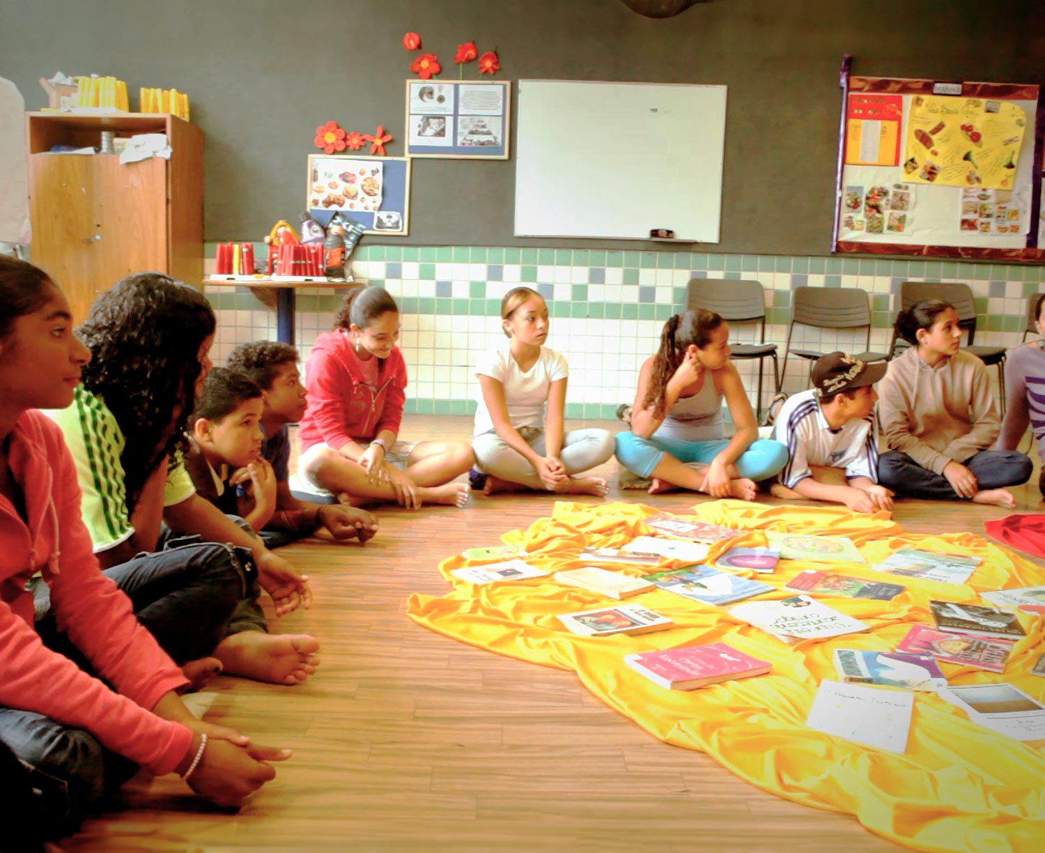 Os alunos de uma escola apoiada pelo fundo Iniciativa Comum no Brasil sentam-se em círculo em uma sala de aula, voltados para a direita, com um cobertor repleto de papéis entre eles.